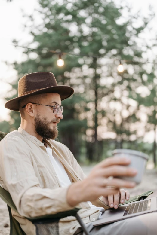 a man sitting in a chair using a laptop, trending on unsplash, renaissance, forest picnic, caracter with brown hat, holds a smart phone in one hand, jewish young man with glasses