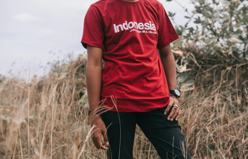 a man standing in a field with a skateboard, inspired by Erik Pevernagie, unsplash contest winner, sumatraism, red t-shirt, ancient indonesia, simple logo, 3/4 front view