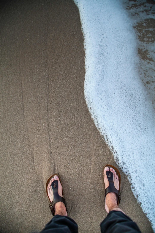 a person standing on a beach next to the ocean, yinyang shaped, sandals, foamy waves, from above