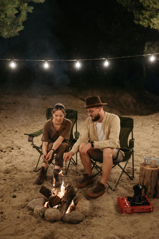 a man and a woman sitting around a campfire, on the sand, string lights, wearing adventure gear, traveller