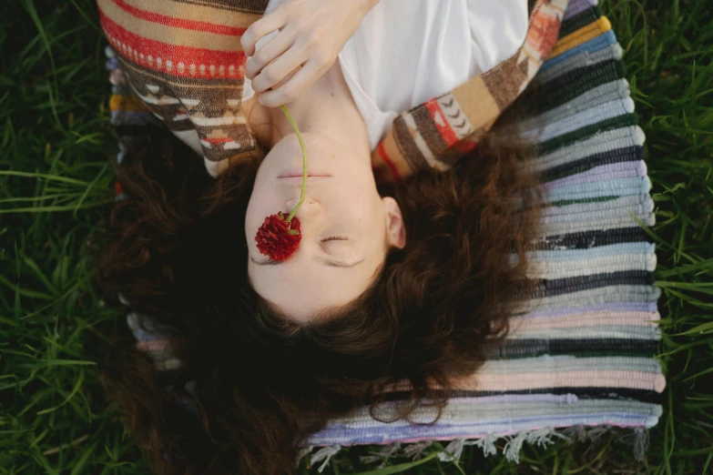 a woman laying on a blanket in the grass, inspired by Elsa Bleda, pexels contest winner, renaissance, giant rose flower as a head, brown curly hair, taking from above, close-up portrait film still