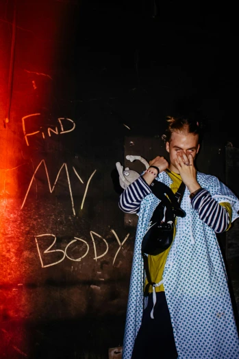 a man standing in front of a wall with writing on it, an album cover, by Anna Findlay, trending on pexels, graffiti, in a nightclub, clothed holy body, frank dillane, looking for clues