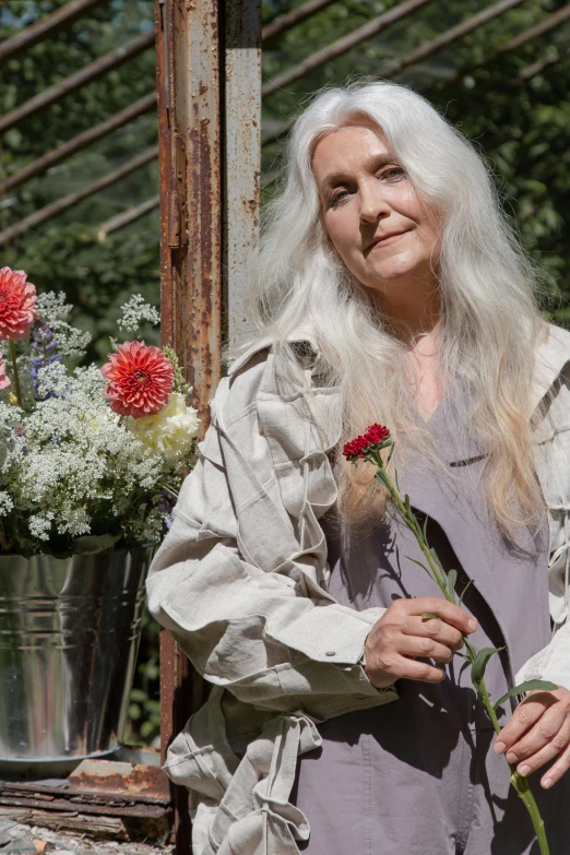 a woman standing in front of a bunch of flowers, a portrait, inspired by Grethe Jürgens, pexels contest winner, light gray long hair, annie lebowitz, promotional image, sitting down