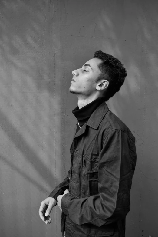 a black and white photo of a man standing in front of a wall, inspired by Jean Malouel, wearing a turtleneck and jacket, bella poarch, head tilted down, sitting down casually