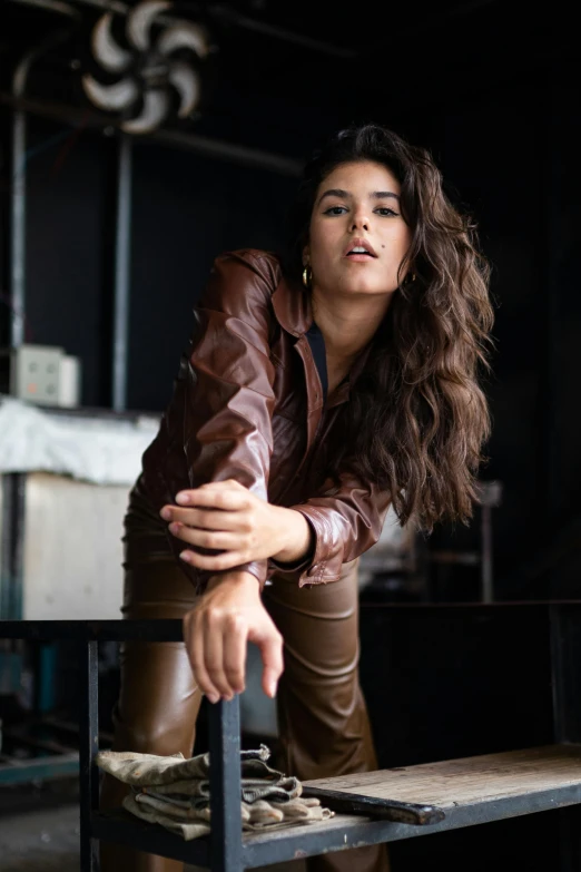 a woman in a leather jacket leaning on a bench, by Robbie Trevino, renaissance, wavy hair spread out, sitting on a mocha-colored table, in an action pose, leather pants | natural lighting