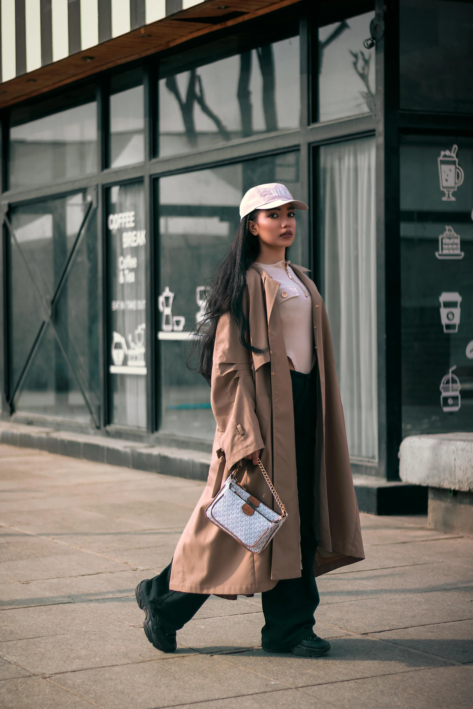 a woman walking down a sidewalk next to a building, pexels contest winner, wearing a long beige trench coat, jakarta, baggy clothing and hat, milk and mocha style