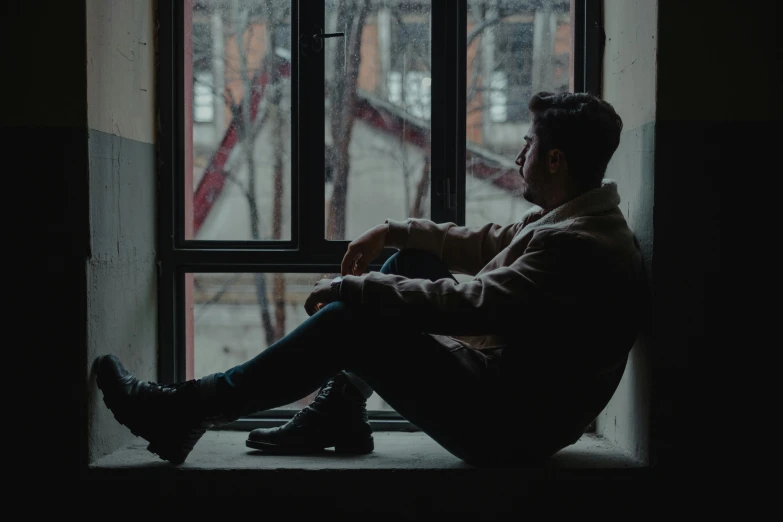 a man sitting on a window sill looking out a window, non-binary, heartbroken, sitting down casually, faded and dusty
