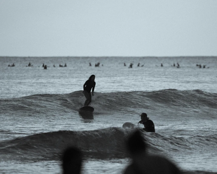 a man riding a wave on top of a surfboard, a black and white photo, pexels contest winner, crowded silhouettes, late evening, people watching around, blurry footage