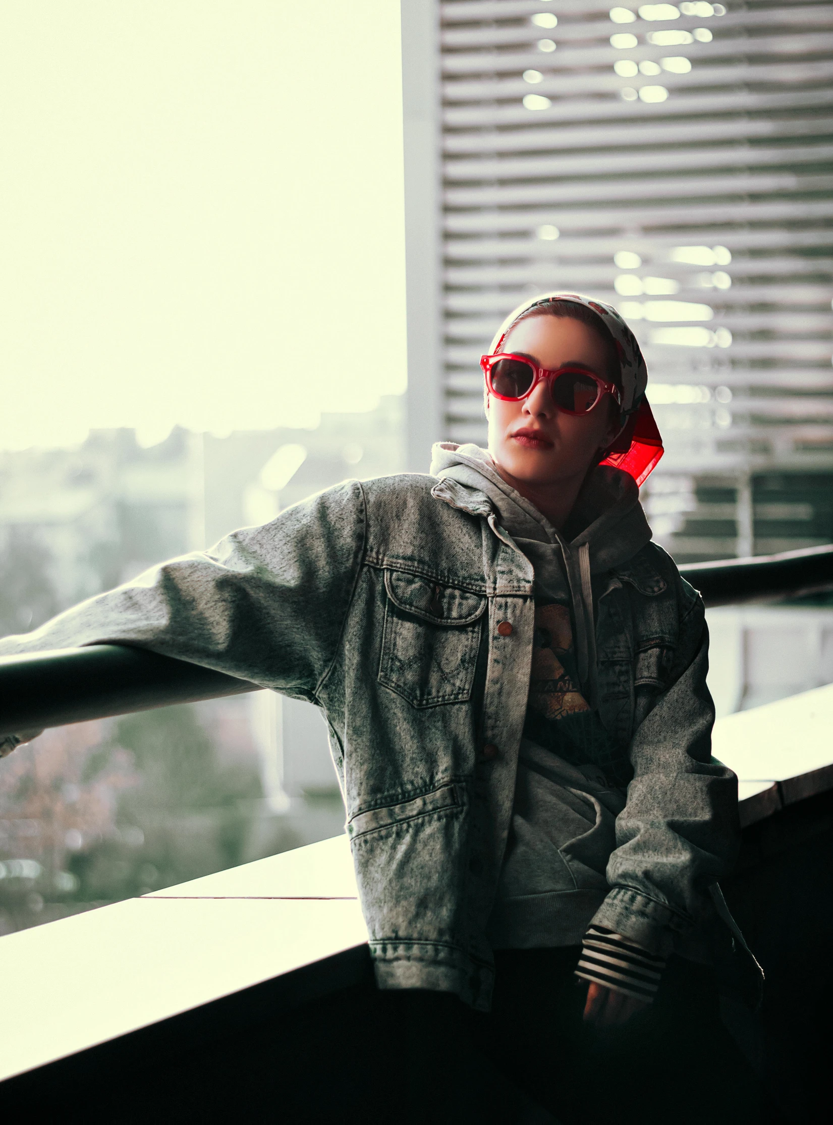 a woman sitting on top of a balcony next to a window, inspired by Elsa Bleda, trending on pexels, wearing red tainted glasses, jean jacket, streetwear, with a red halo over her head