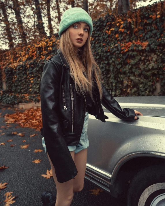 a woman standing next to a parked car, a colorized photo, inspired by Elsa Bleda, trending on pexels, black beanie, she wears leather jacket, ((greenish blue tones)), autum