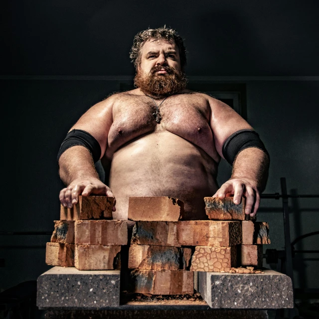 a man sitting on top of a pile of bricks, big beard, wwe, bulky build, press shot