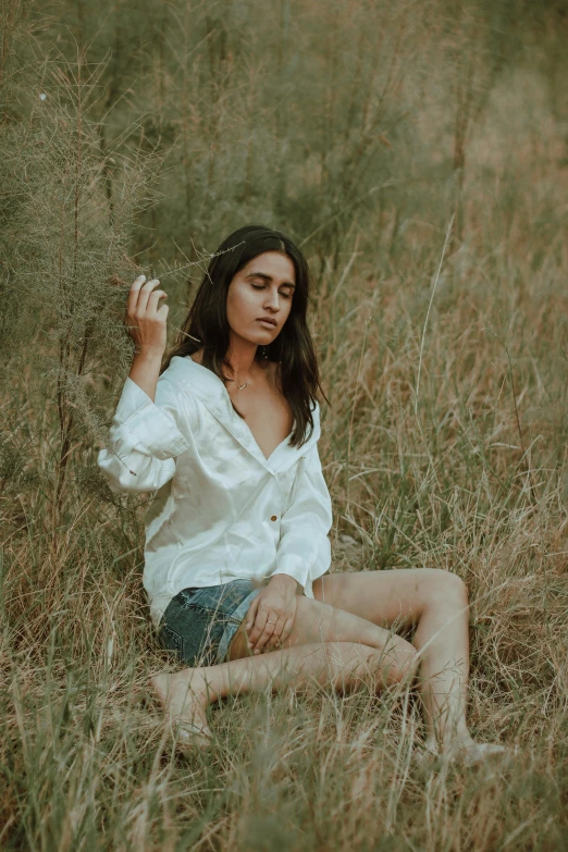 a woman sitting in a field holding a cigarette, inspired by Elsa Bleda, pexels contest winner, wearing a white button up shirt, wearing a camisole and shorts, provocative indian, grass
