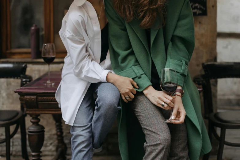 a couple of women sitting next to each other, by Emma Andijewska, trending on pexels, green corduroy pants, trench coat and suit, romantic couple, with a drink