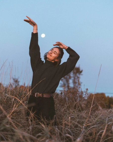 a woman standing on top of a grass covered field, with the moon out, non binary model, she is dancing, wearing a turtleneck and jacket