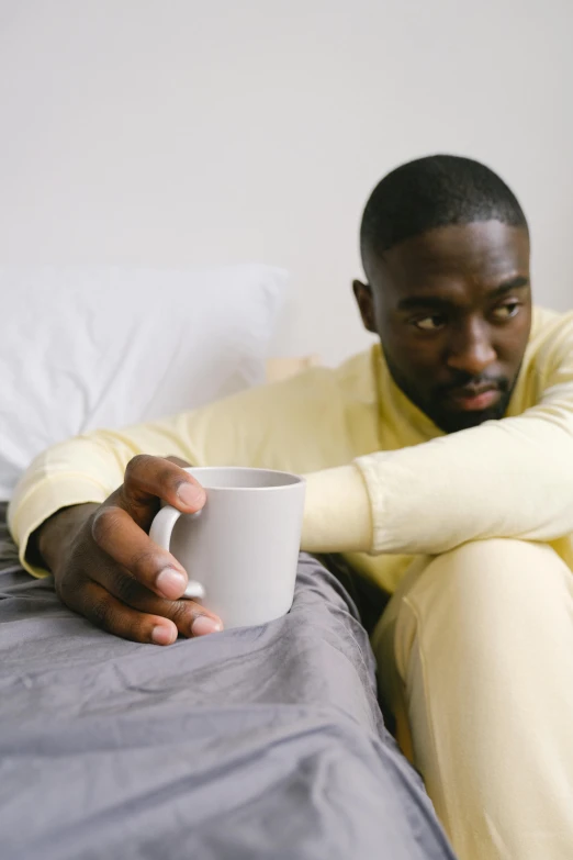 a man sitting on a bed holding a cup, by Stokely Webster, disappointed, yellow, cup of coffee, wearing pajamas