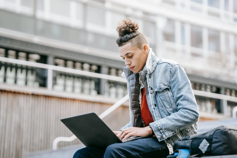 a man sitting on a bench using a laptop, trending on pexels, woman with braided brown hair, aboriginal australian hipster, non-binary, wearing a jeans jackets