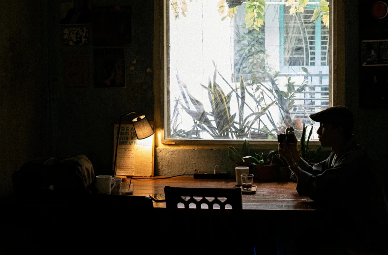 a person sitting at a table in front of a window, inspired by Elsa Bleda, unsplash contest winner, lantern light besides, cuban setting, small hipster coffee shop, two buddies sitting in a room