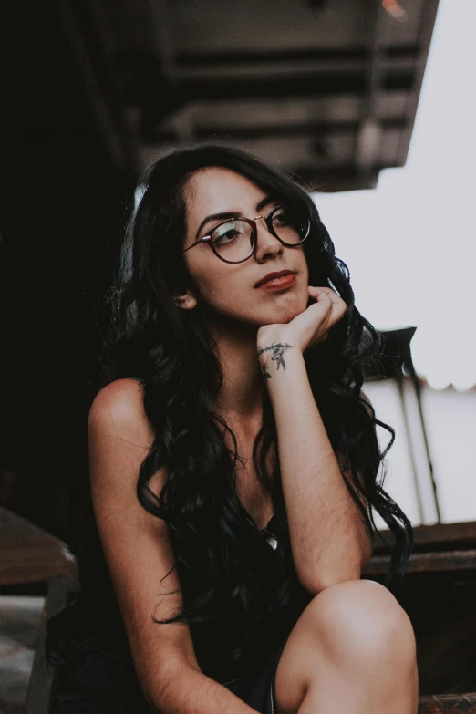 a woman sitting on the steps of a building, by Robbie Trevino, trending on pexels, wavy long black hair and glasses, with tattoos, discord profile picture, profile image