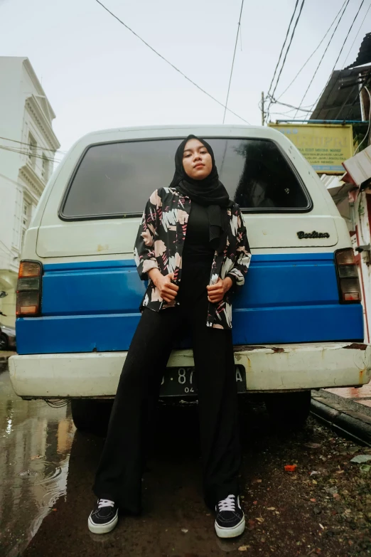 a woman standing in front of a blue and white van, an album cover, by Basuki Abdullah, wearing a fancy black jacket, ((portrait)), high quality upload, vine