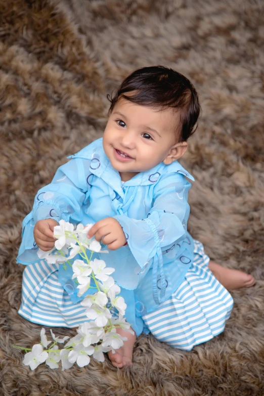 a baby sitting on top of a rug holding a flower, an album cover, inspired by Nazmi Ziya Güran, shutterstock contest winner, icey blue dress, cute young man, high resolution photo, blue colored traditional wear
