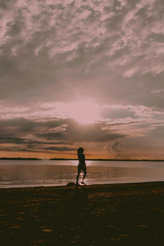 a person standing on a beach next to a body of water, pexels contest winner, girl clouds, light over boy, low quality photo, low sun