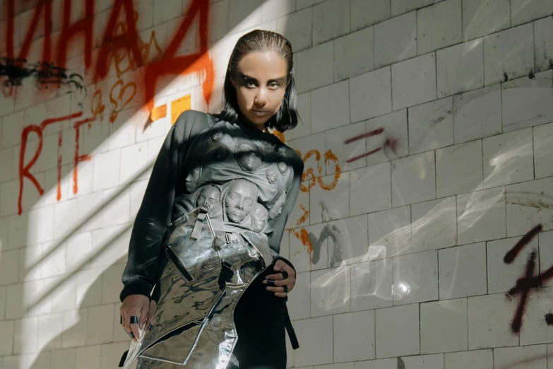 a woman standing in front of a wall with graffiti on it, an album cover, inspired by Anna Füssli, wearing techwear and armor, frank dillane, official store photo, silver garment