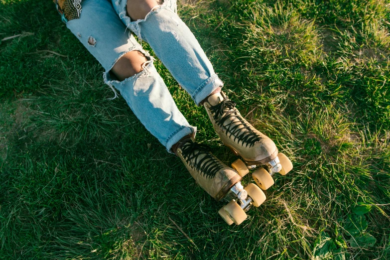 a person sitting on a skateboard in the grass, jeans and boots, brass wheels, tan, viewed from the ground