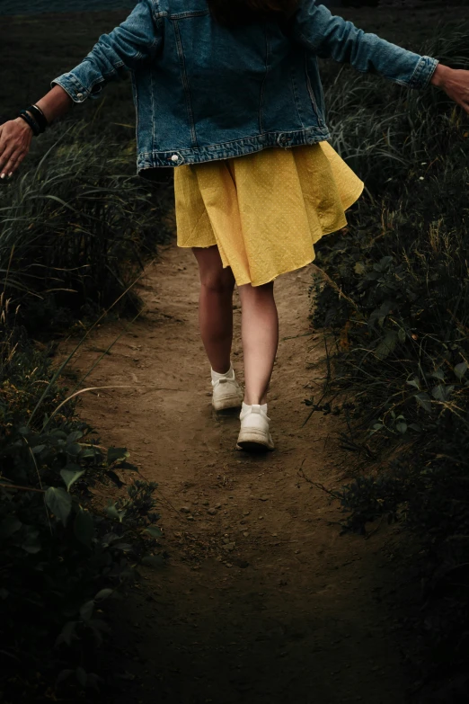 a woman walking down a dirt path with her arms outstretched, by Lucia Peka, pexels contest winner, yellow clothes, wearing skirt, young girl, legs intertwined