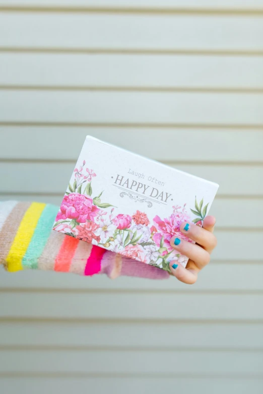 a woman holding a card in her hand, inspired by Eden Box, happy color, beautiful day, full product shot, pink flowers