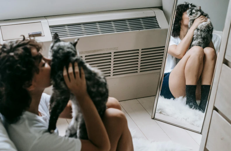 a woman sitting in front of a mirror holding a cat, air conditioner, background image, hugging her knees, portrait featured on unsplash