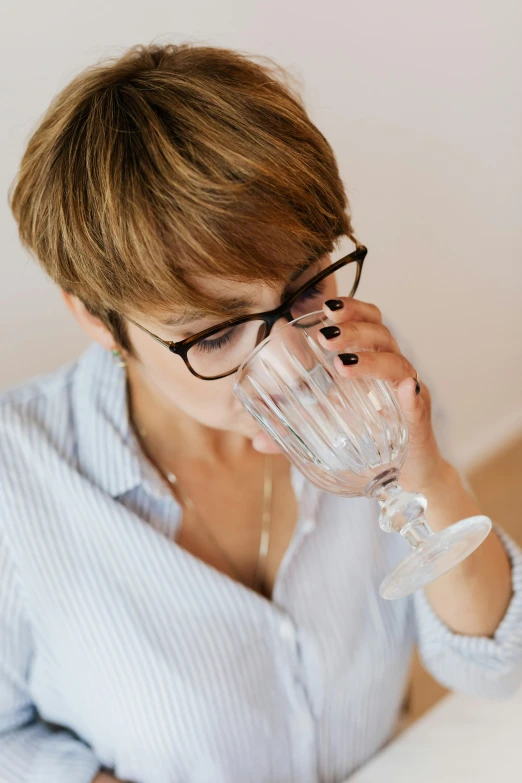 a woman sitting at a table drinking from a glass, black rimmed glasses, water to waste, well defined, sleepy