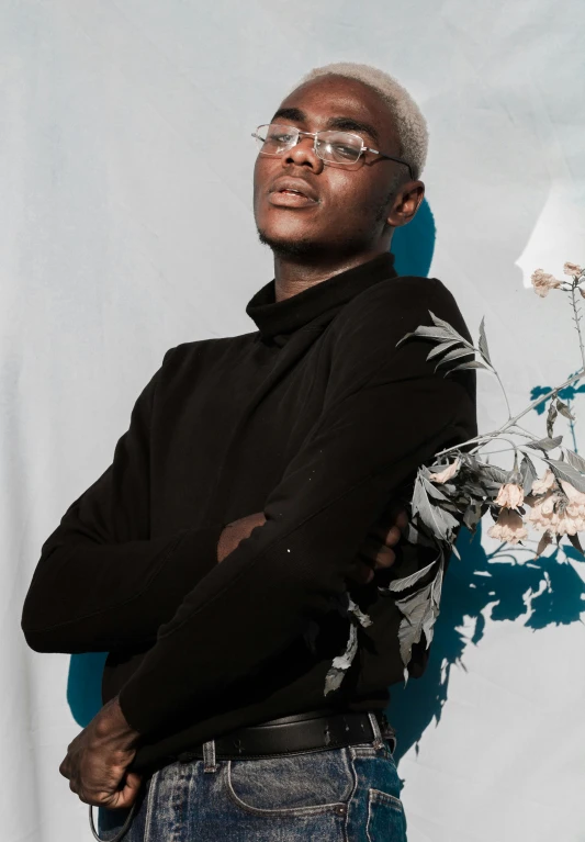 a man standing in front of a blue wall, an album cover, inspired by Camille-Pierre Pambu Bodo, trending on unsplash, afrofuturism, silver haired, flowers growing out of his body, black turtleneck, black teenage boy