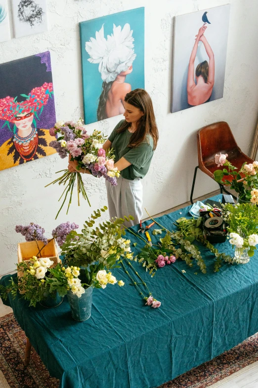 a woman arranging flowers on a table in a room, process art, teal studio backdrop, market setting, overview, tall