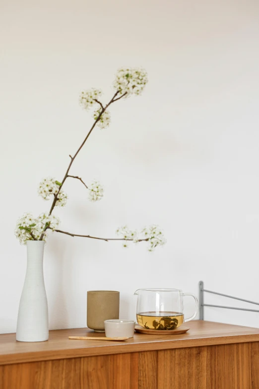 a white vase sitting on top of a wooden dresser, a still life, inspired by Kanō Shōsenin, minimalism, iced tea glass, almond blossom, at a dinner table, white backround