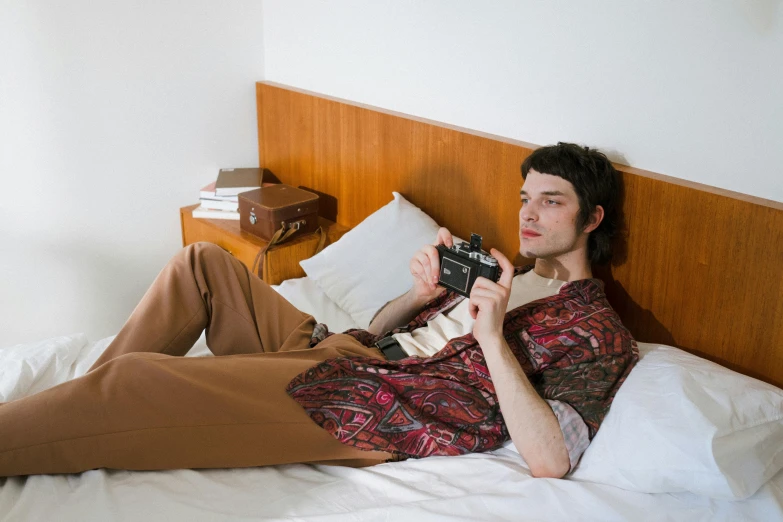 a man laying on a bed holding a camera, inspired by Balthus, visual art, dressed in 1970s menswear, paisley, brown pants, jonny greenwood