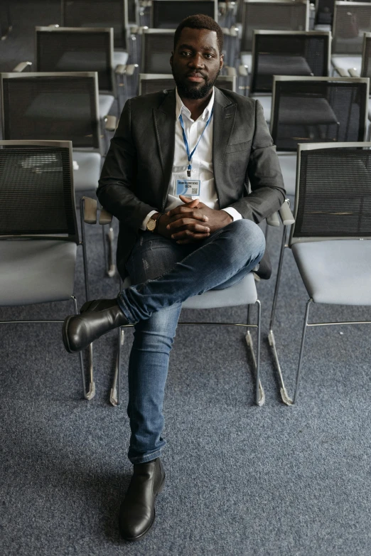 a man sitting on a chair in a room full of chairs, dark skinned, ready for a meeting, crossed legs, full body hero