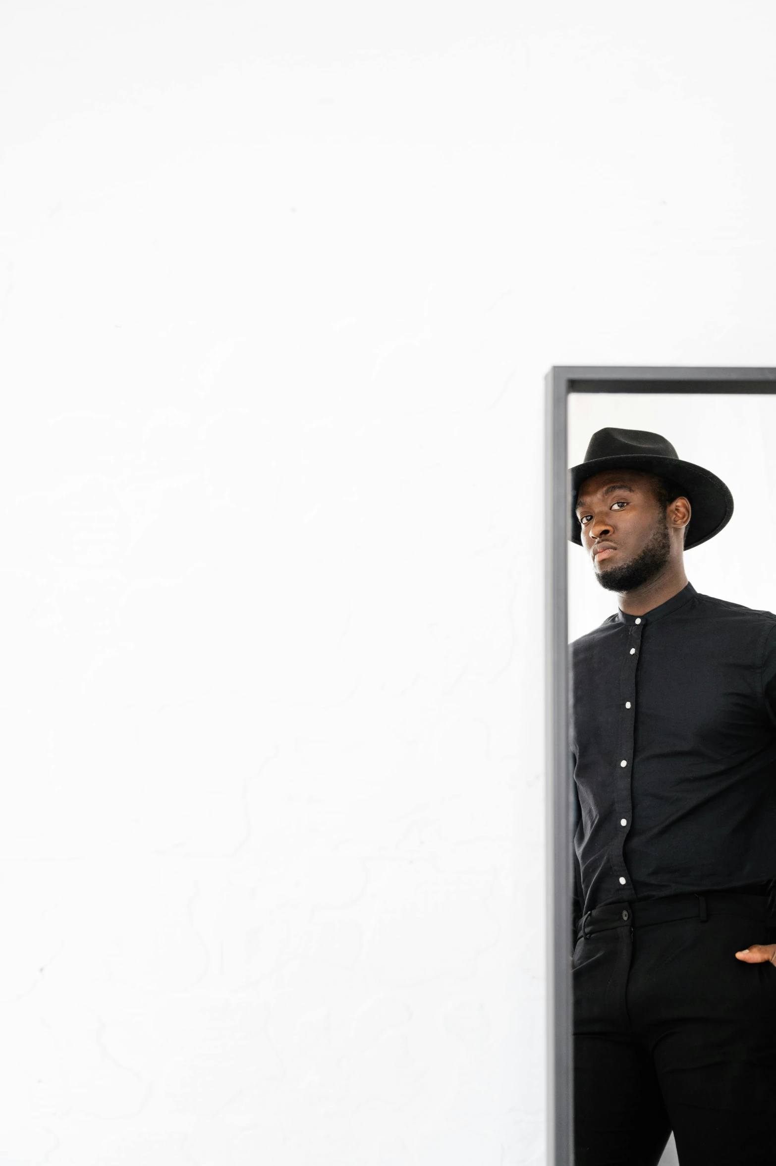 a man in a hat standing in front of a mirror, an album cover, by Nina Hamnett, pexels contest winner, minimalism, sharp black skin, in black uniform, leaning against the wall, dwell