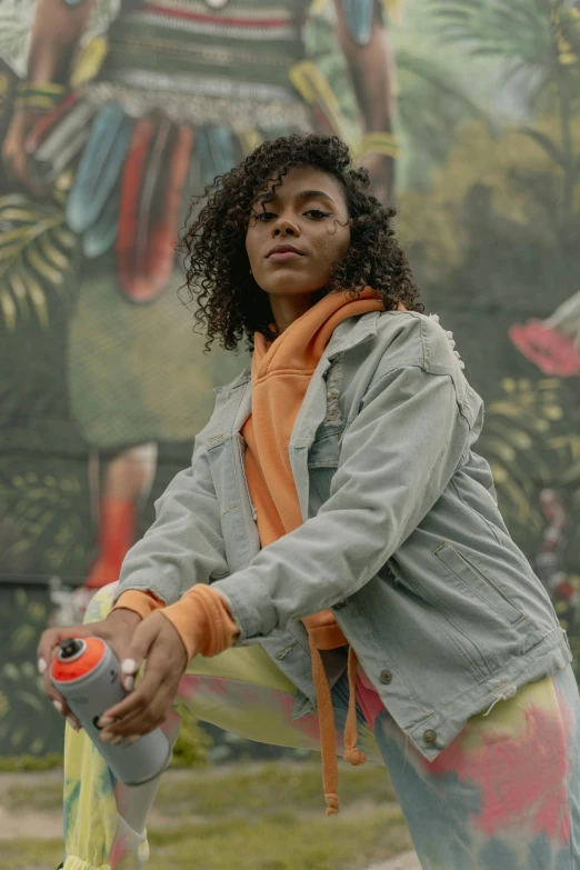 a woman holding a tennis racquet in front of a mural, an album cover, trending on unsplash, graffiti, orange jacket, tessa thompson, casual pose, juice