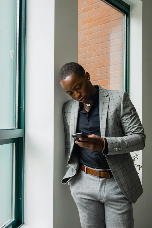 a man standing by a window looking at his cell phone, wearing dark grey suit, dark skin tone, schools, mobile app