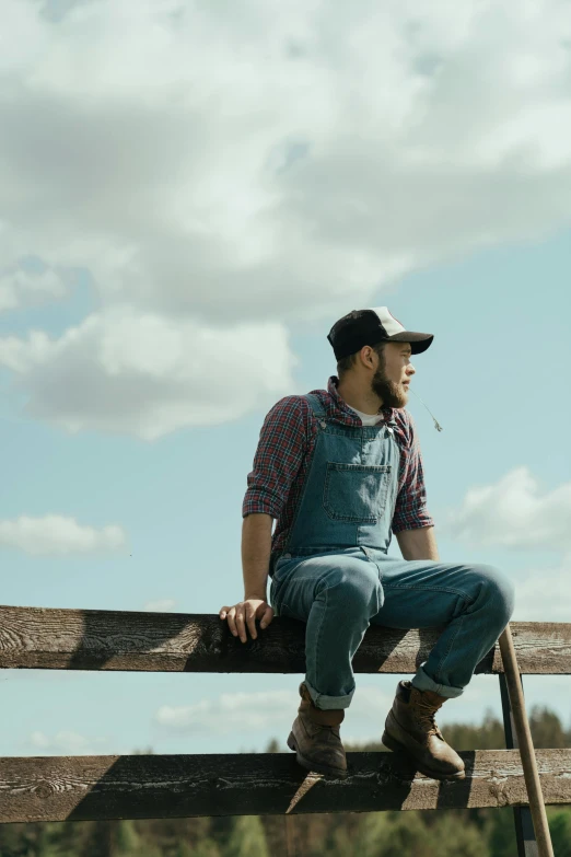 a man sitting on top of a wooden fence, wearing farm clothes, looking at the sky, wearing overalls, zachary corzine
