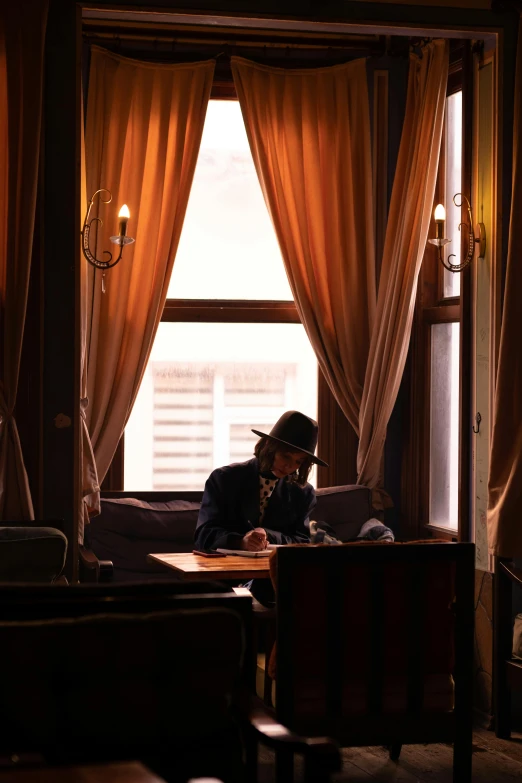 a man sitting at a table in front of a window, inspired by Brassaï, unsplash, caracter with brown hat, gentleman's club lounge, writing a letter, high quality photo