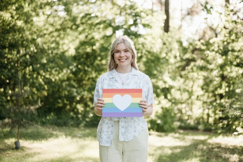 a woman standing in a field holding a rainbow heart sign, by Alice Mason, pexels, in front of a forest background, official print, rectangle, kalevala