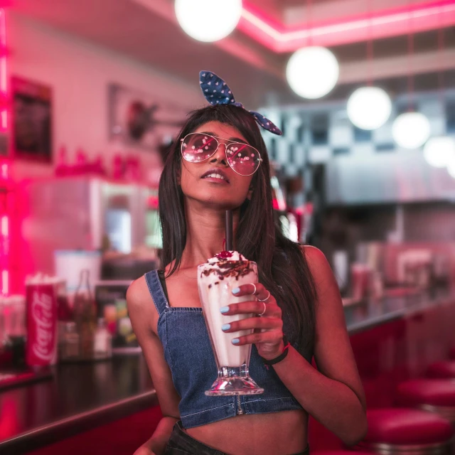 a woman sitting at a bar holding a glass of milkshake, trending on pexels, pop art, brandon woelfel, pink glasses, indian girl with brown skin, 5 0's diner