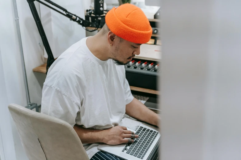 a man sitting in front of a laptop computer, an album cover, trending on pexels, wearing an orange t shirt, beanie, news broadcast, kyoto studio
