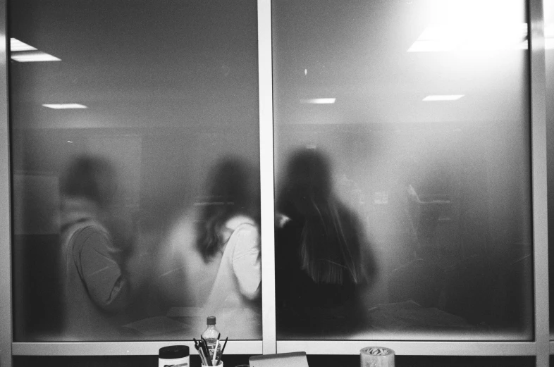 a couple of people standing in front of a window, a black and white photo, by Emma Andijewska, unsplash, process art, sitting in office, ghost faces, view from behind mirror, morning haze