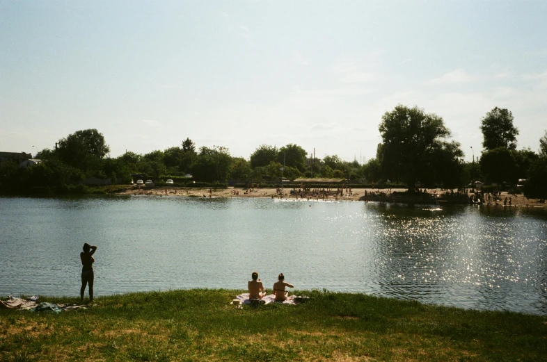 a group of people standing on top of a lush green field, a photo of a lake on a sunny day, kreuzberg, sandy beach, people swimming