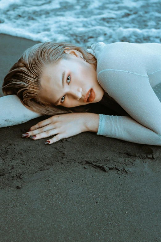 a woman laying on a beach next to the ocean, inspired by Elsa Bleda, unsplash, surrealism, blonde hair and blue eyes, portrait sophie mudd, on a gray background, androgynous person