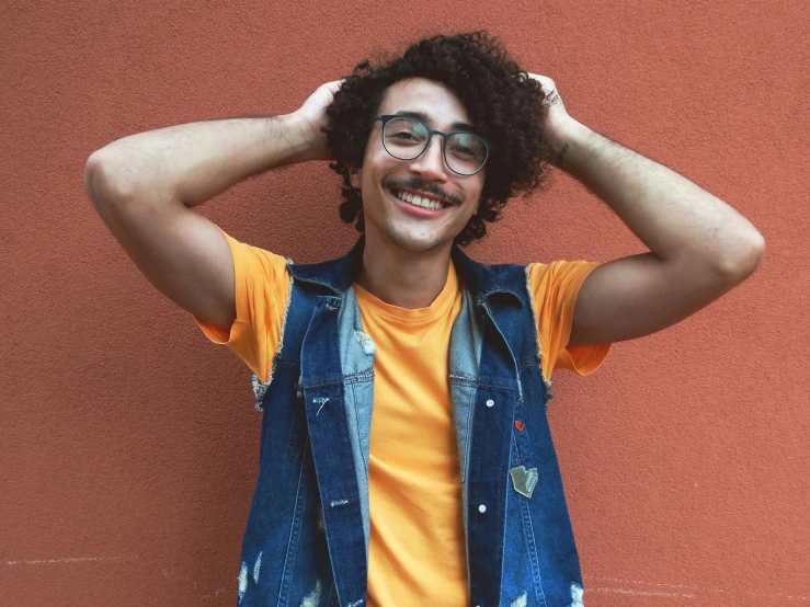 a man standing in front of a wall with his hands on his head, an album cover, pexels, antipodeans, cute slightly nerdy smile, yellow-orange, dark short curly hair smiling, raden saleh