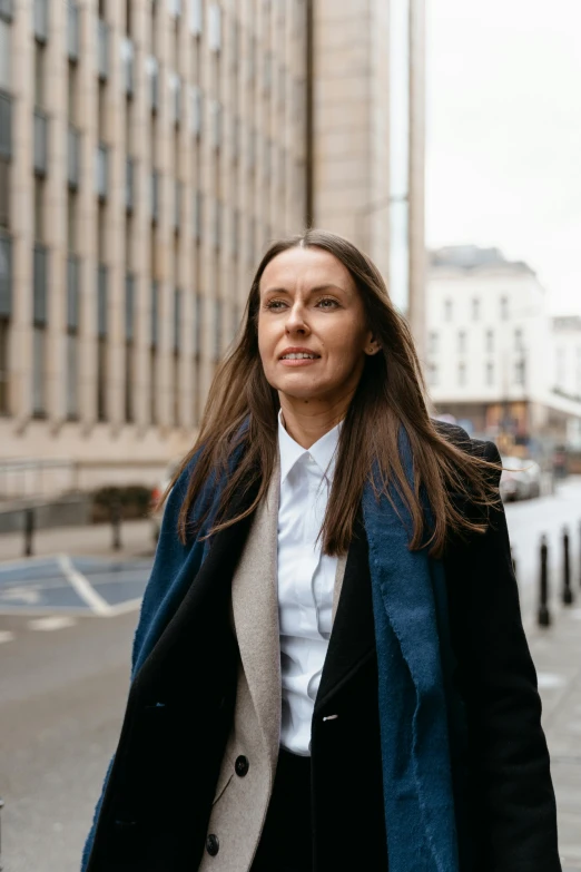 a woman walking down a street next to tall buildings, inspired by Jemima Blackburn, pexels contest winner, wearing lab coat and a blouse, liz truss, wearing jacket, female with long black hair