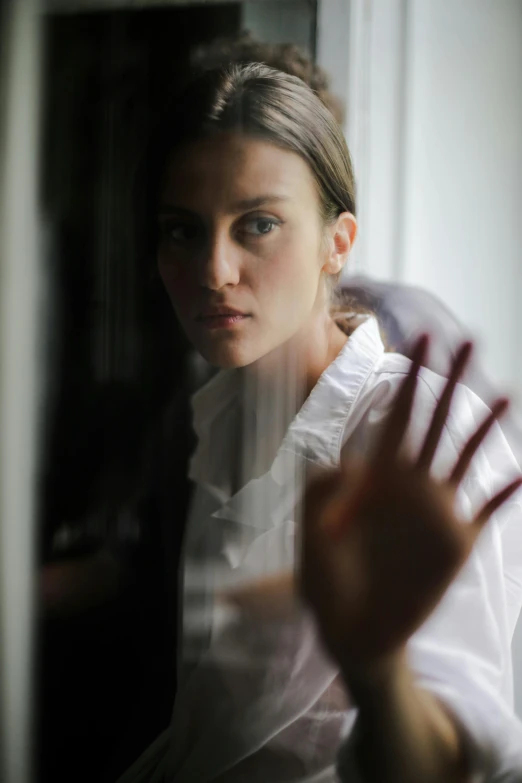 a woman in a white shirt is looking out a window, creating an ominous presence, looking in a mirror, promo image, solemn gesture